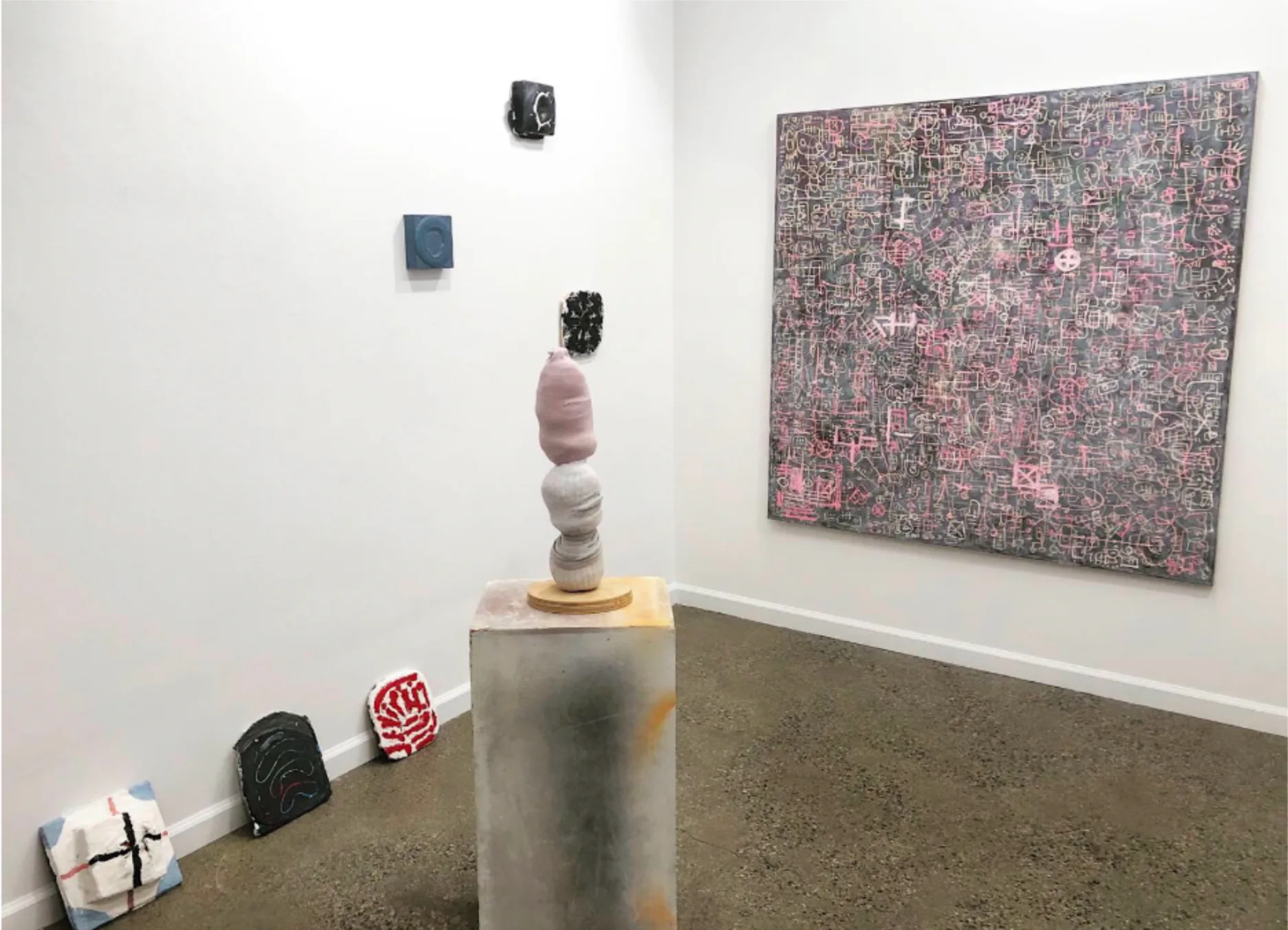 Gallery installation with stacked sculptural objects on pedestal, wall-mounted abstract artwork, and objects on floor in contemporary art space.