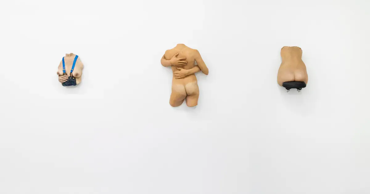 Three small wooden figurative sculptures in natural tan wood displayed in a row against white wall: an abstracted head form, a kneeling torso, and a horizontal abstraction.
