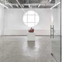 Minimal gallery interior with circular skylight, polished concrete floor, glass barriers, and sparse sculptural artwork on pedestal.