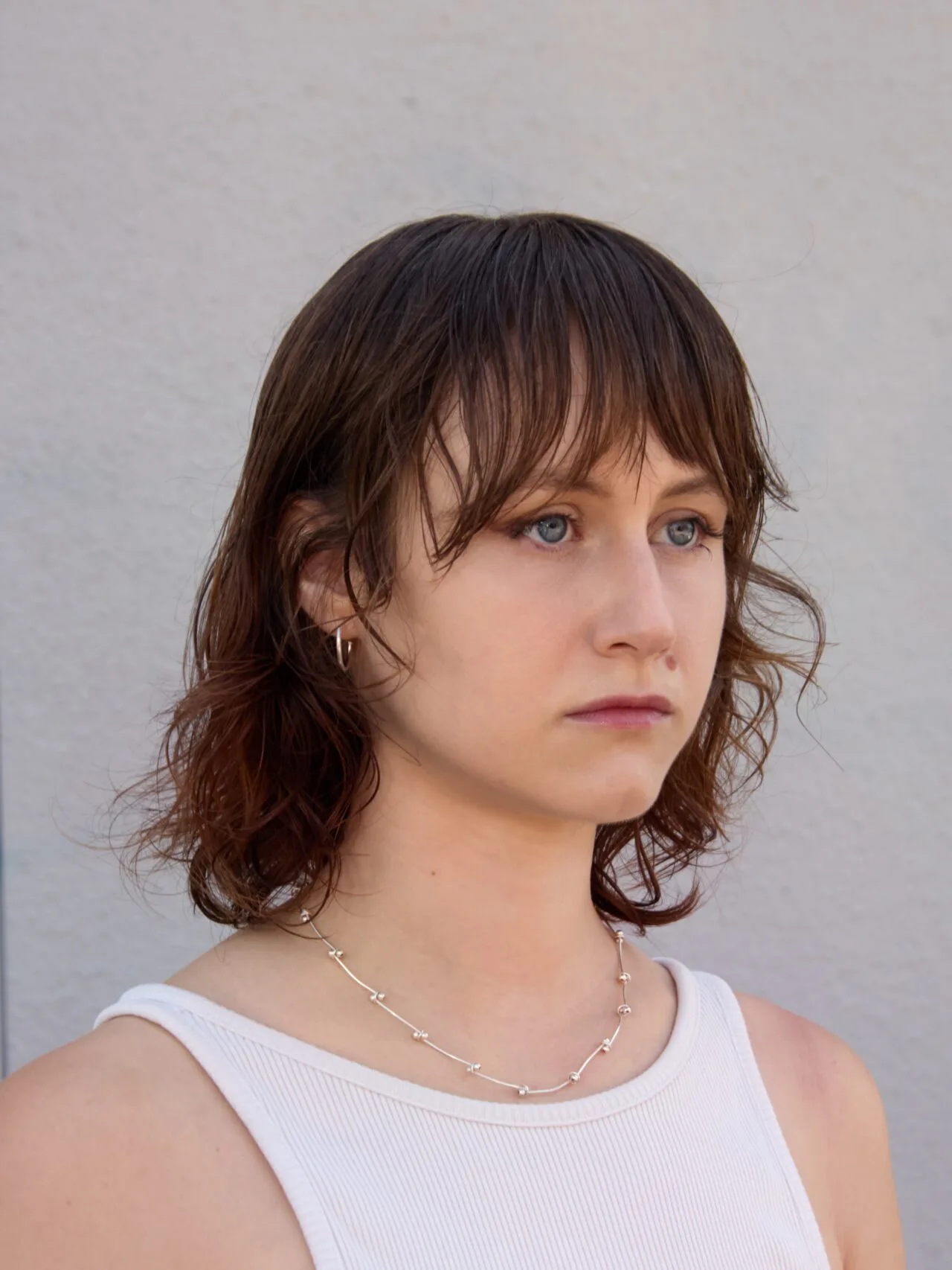 Person with shoulder-length reddish-brown wavy hair and bangs, wearing a white sleeveless top and silver chain necklace, looking slightly to the side.