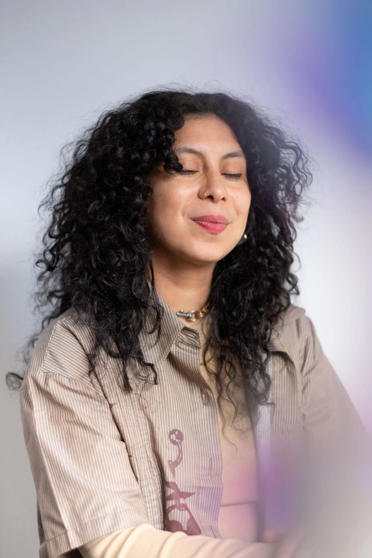 Person with long curly black hair, eyes closed, wearing pink lipstick and a beige ribbed long-sleeve shirt, against purple-tinted gradient background.
