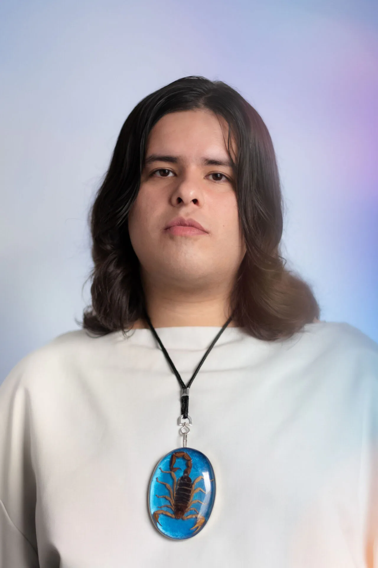 Young man with long dark wavy hair wearing a white shirt and a scorpion pendant necklace, facing the camera against a soft purple-blue backdrop