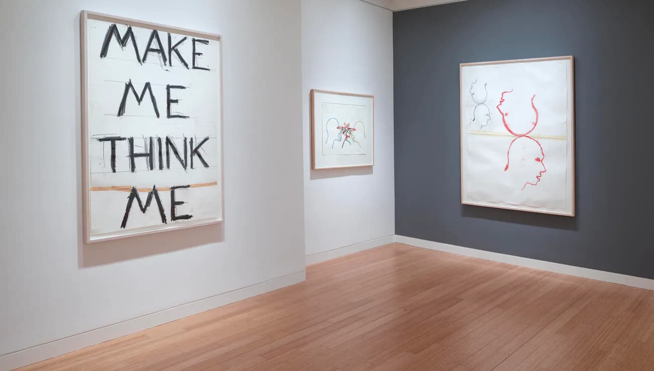 Gallery interior with wooden floors, white and dark gray walls displaying framed text-based and abstract artworks.
