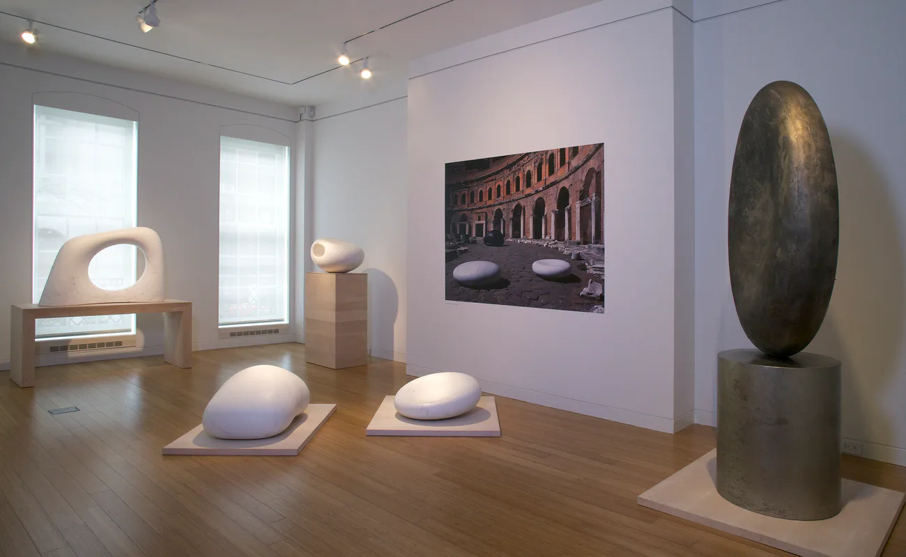 Gallery with wooden floors featuring smooth curved sculptural forms on pedestals, large dark metallic oval sculpture, and architectural photograph.