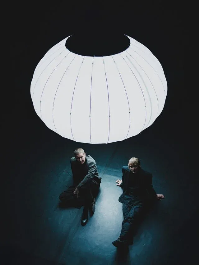 Two figures sit beneath a large white inflated sculptural form with vertical ribbing against dark teal background.