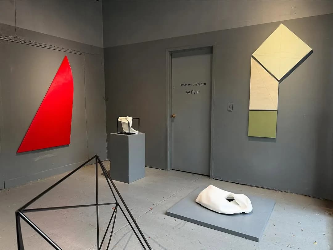 Gallery interior featuring a red geometric wall installation, yellow-white angular wall piece, metal geometric sculpture, and white dome-shaped artwork on concrete floor.