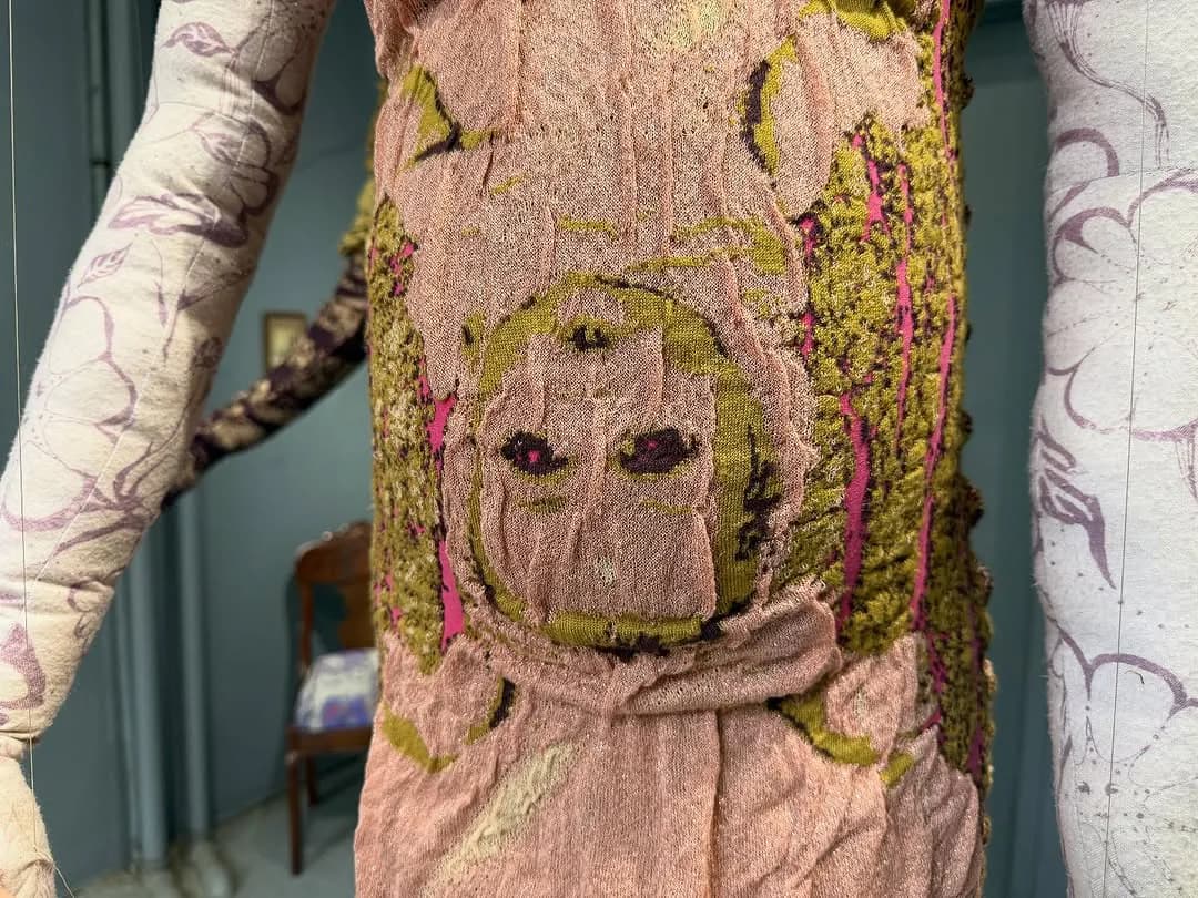 Close-up textile artwork featuring an embroidered or stitched face on burlap with moss-green and pink thread details on natural fabric.