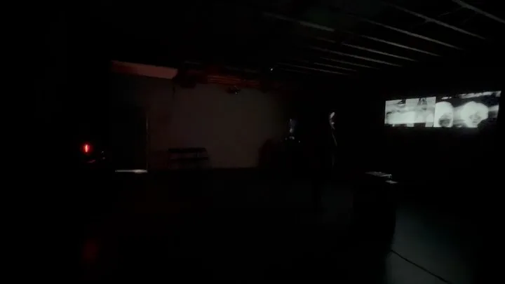 Dark gallery interior with minimal lighting, a red light indicator visible, and a video projection on the wall in distance.