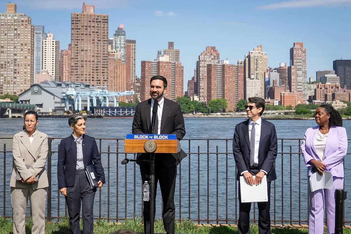 Five people stand at a podium overlooking a waterfront with Manhattan skyline in background; urban waterfront venue with metal railing.