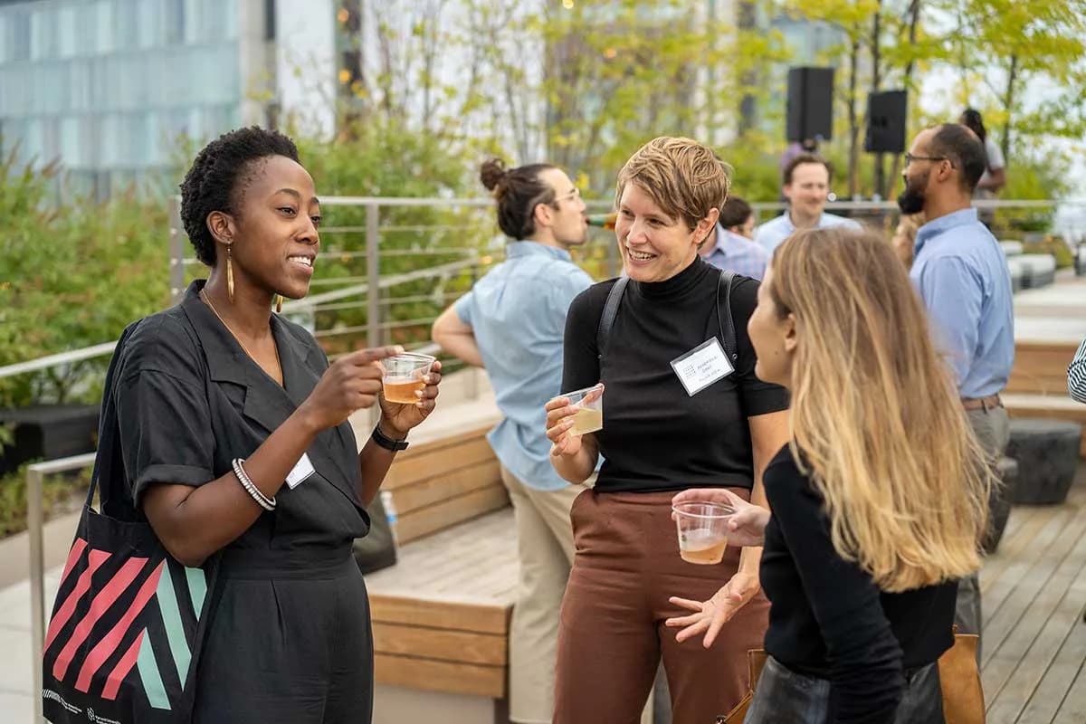 People socializing with drinks on an outdoor rooftop deck with wooden seating and modern building in background.