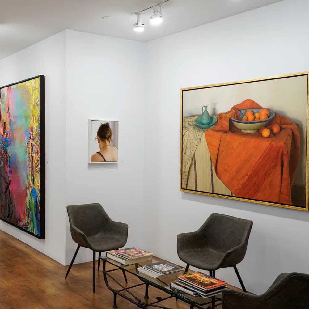 Gallery interior with white walls, track lighting, wood floors, gray armchairs, and paintings including an abstract piece, a portrait, and a still life of oranges