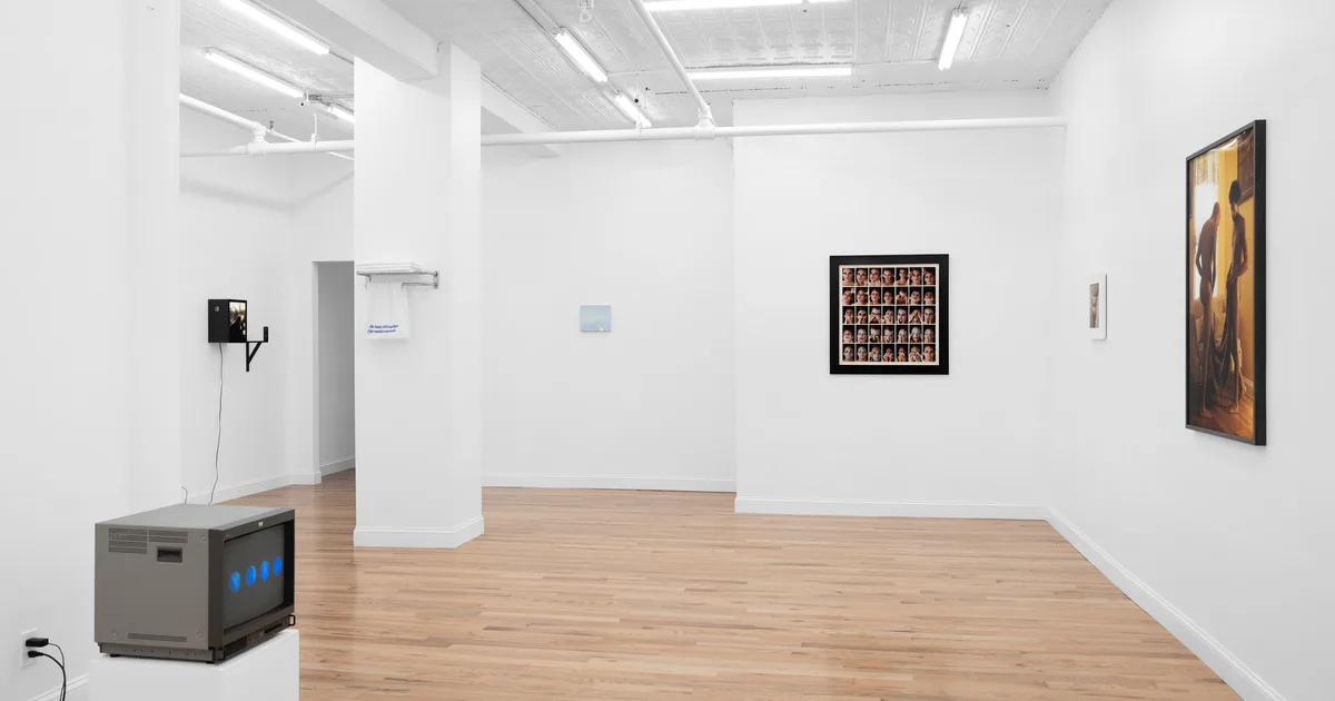 Gallery interior with white walls, wood flooring, and framed artworks displayed alongside a video monitor sculpture