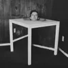 Black and white photograph of a minimalist white table with tapered legs in a sparse interior setting.