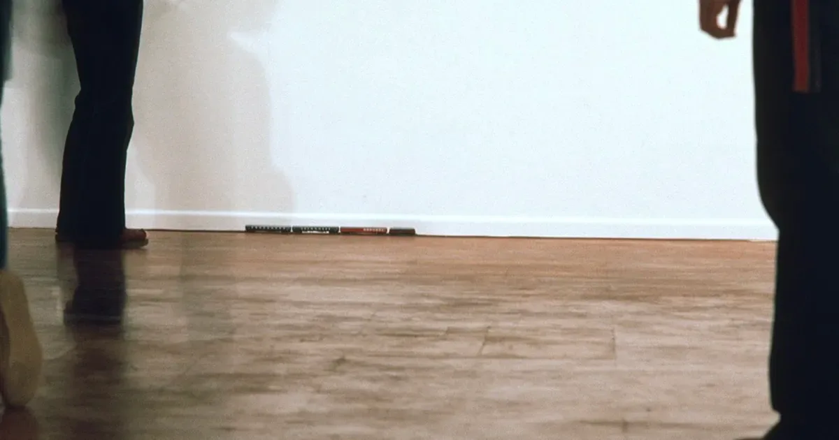 Gallery interior view of wooden flooring with two figures standing in foreground wearing dark clothing, white wall visible in background.
