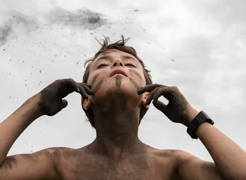 Photograph of shirtless person looking upward with hands framing face against cloudy sky; artistic portrait with textured ground.