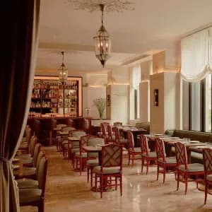 Interior of an elegant gallery or salon room with burgundy chairs, ornate ceiling details, hanging lantern, and tall windows along cream-colored walls.