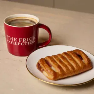 Red ceramic mug printed with 'The Frick Collection' beside a pastry on a white plate on a light surface.