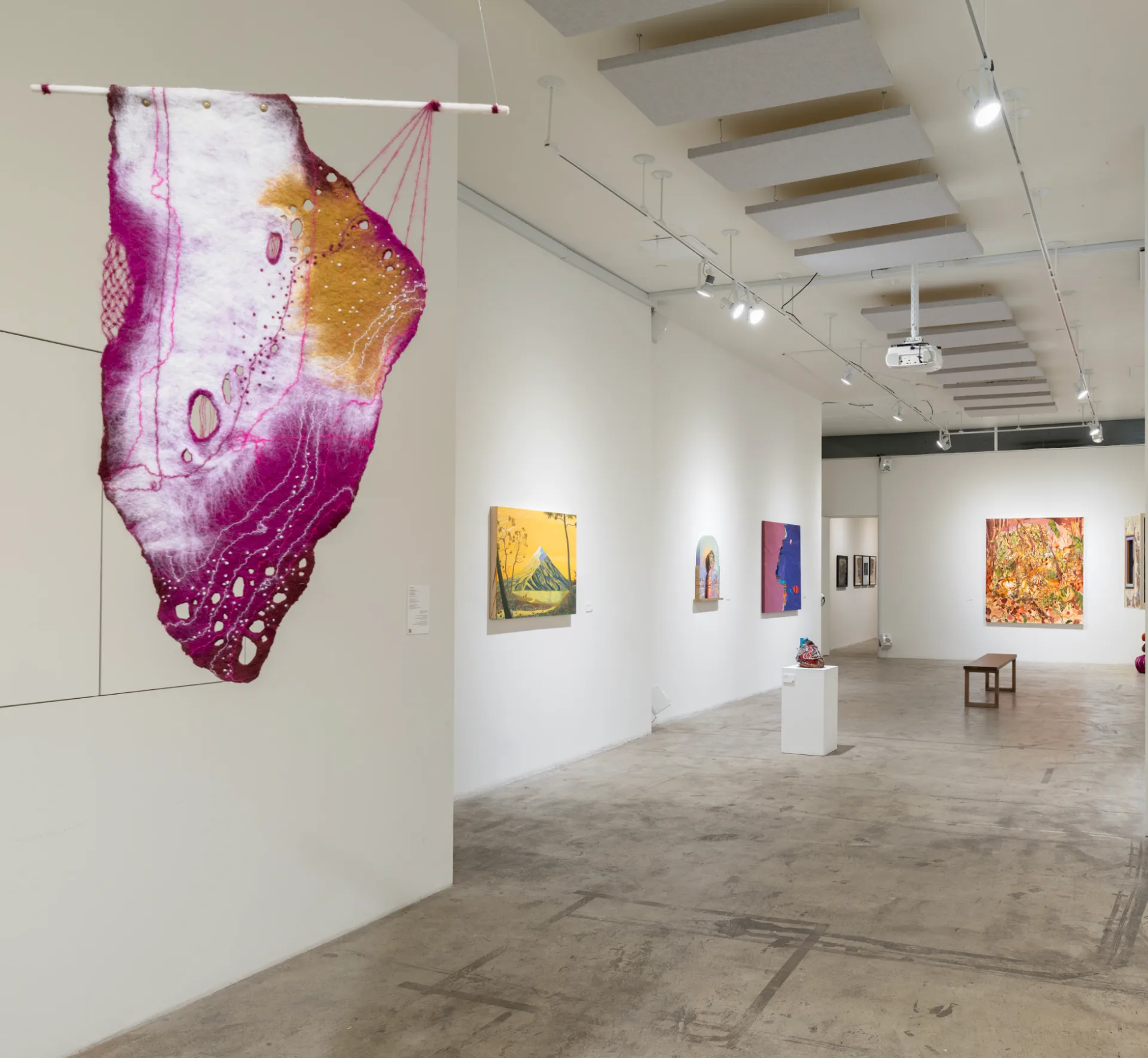 Gallery interior with suspended fuchsia and yellow fabric installation in foreground and multiple artworks visible throughout bright white space.