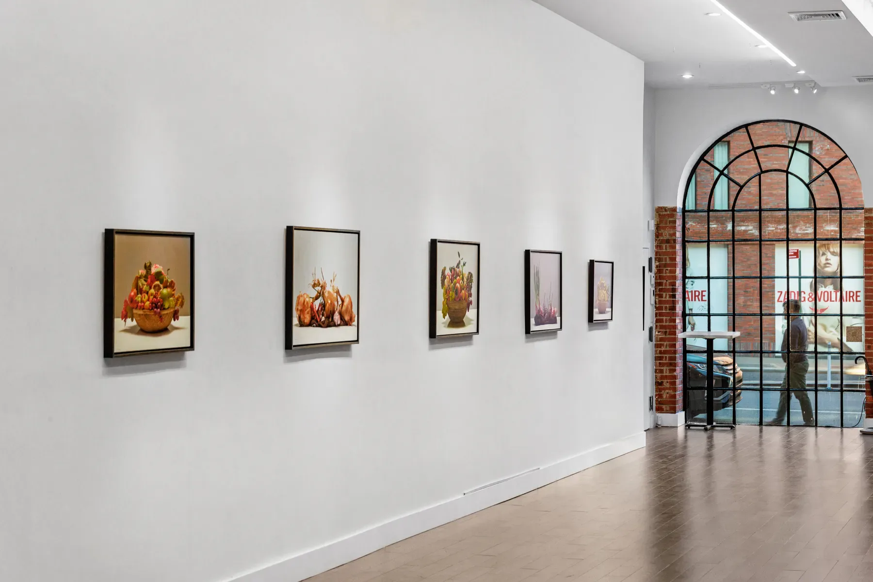 Gallery interior with white walls displaying five framed still-life artworks, wooden floor, and an arched glass entrance leading to the street