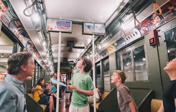 Interior of a vintage subway car with passengers standing and conversing; green and yellow seating visible along the sides.