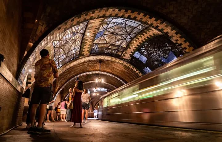 Ornate arched subway station platform with lit architectural details; a train with motion blur passes while passengers wait on the platform.