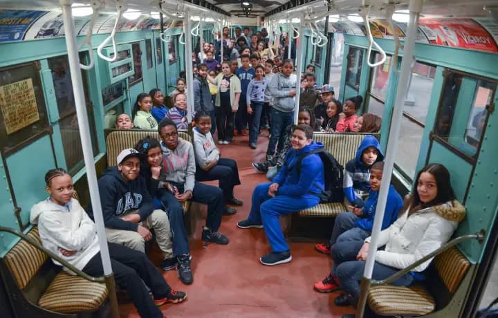 Interior view of a crowded turquoise-and-red vintage subway car filled with seated and standing passengers.