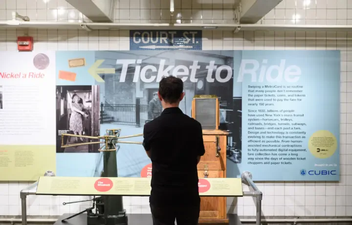 Museum display panel titled "Ticket to Ride" featuring historical transit photography and text, with a person viewing the exhibition.