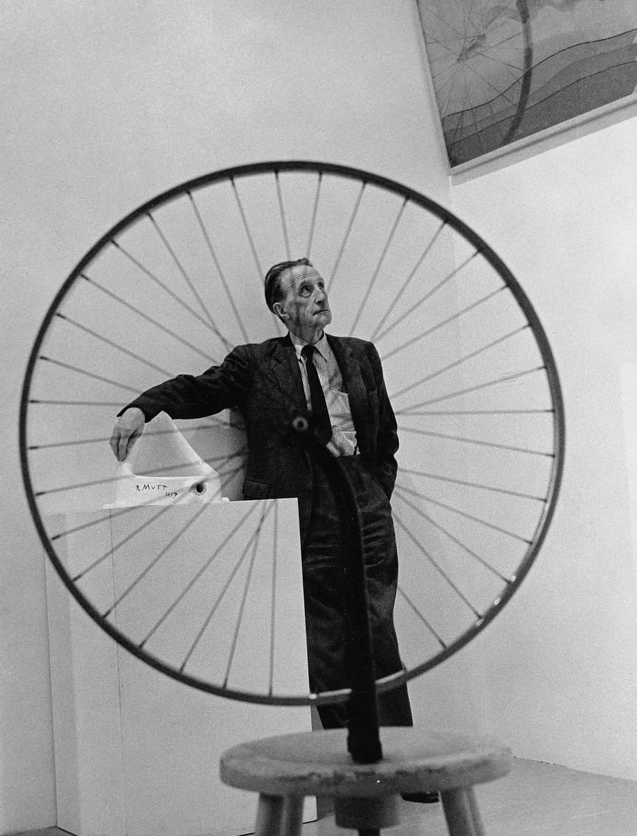 Black and white photograph of a man in suit and tie standing behind a large circular wheel structure, arm extended.