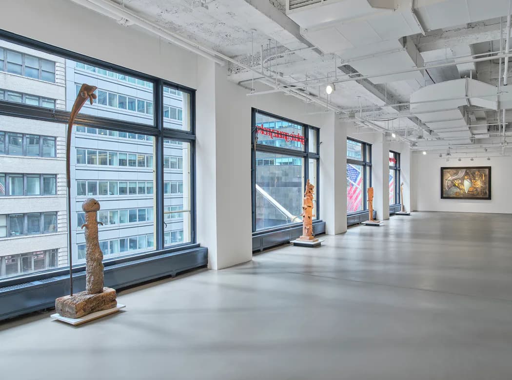Minimalist gallery interior with polished concrete floor, white walls, tall windows overlooking city buildings, and scattered wooden sculptural pieces.