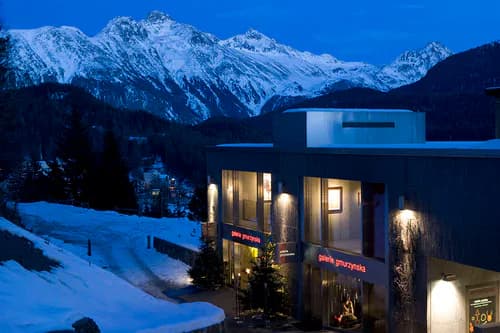 Contemporary white gallery structure with illuminated entrance nestled in snowy alpine landscape at dusk, with snow-covered mountains in background.