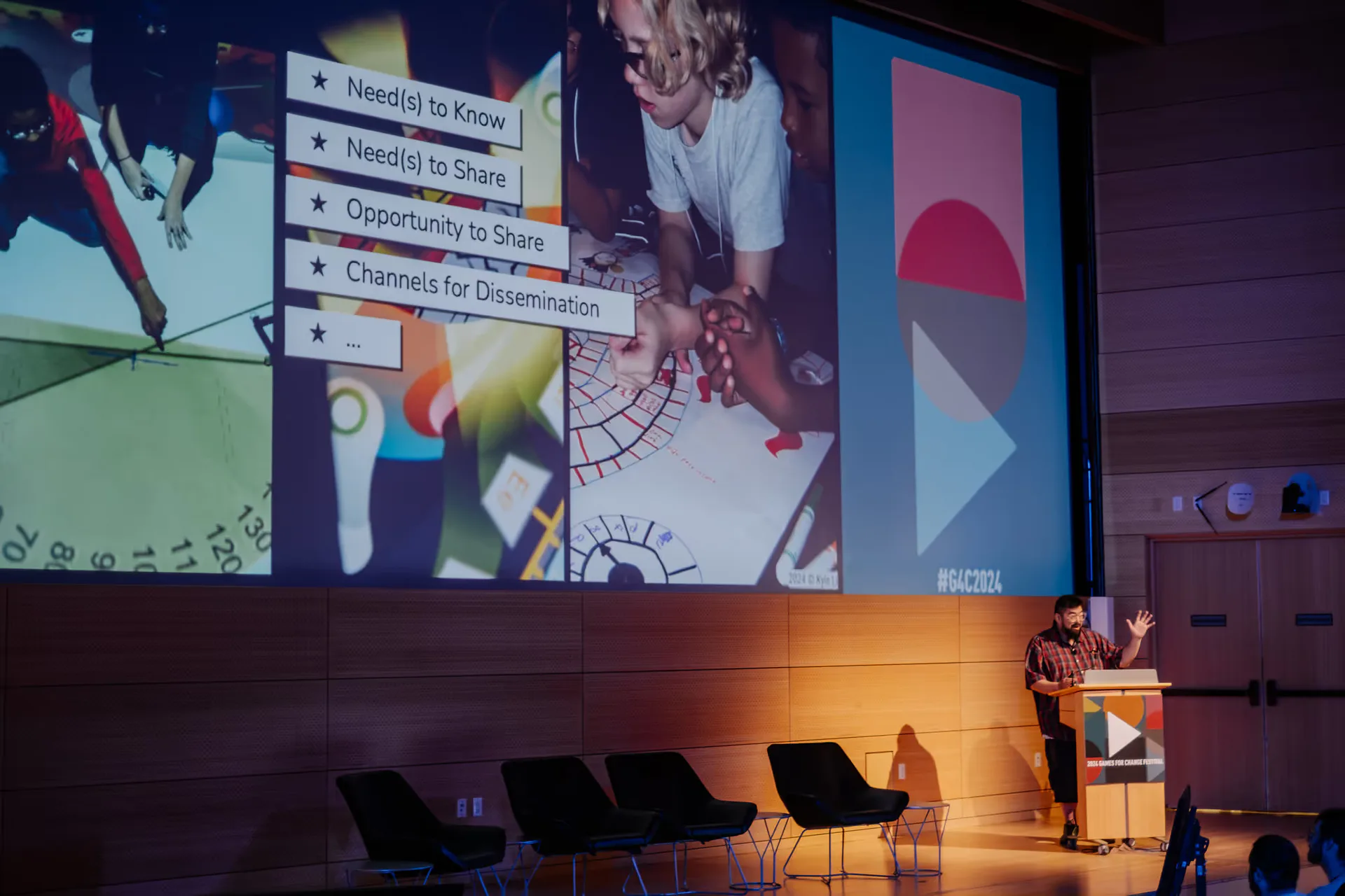 Interior photograph of a presentation space with a speaker at a podium and large projection screen displaying Games for Change content, with audience seating visible.