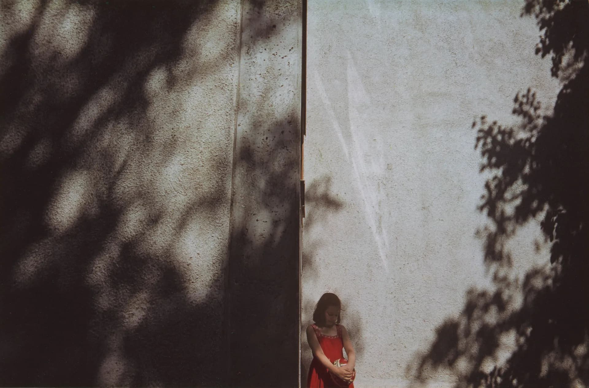 Person in red dress standing against white wall with dappled shadows from foliage cast across the surfaces.