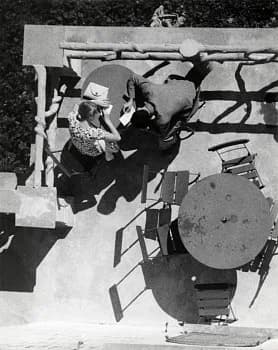 Overhead black and white photograph of person surrounded by chairs and circular objects, compositional arrangement.