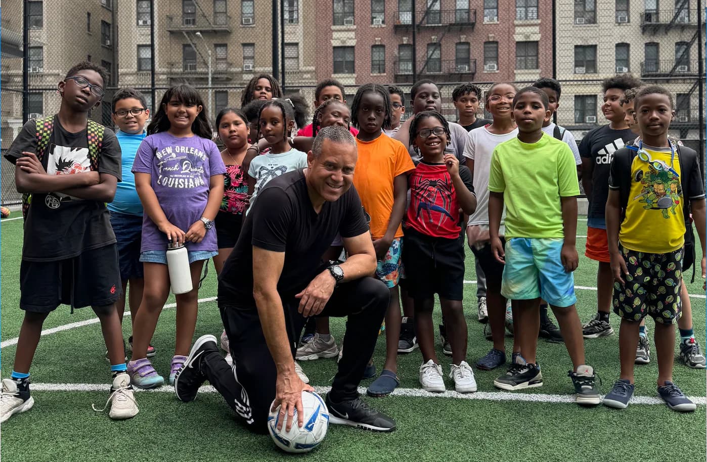 Adult male coach in center crouching with soccer ball surrounded by diverse group of youth players on artificial turf field.
