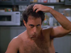 Shirtless man with dark hair touching his head with one hand, photographed indoors against a blurred kitchen background.