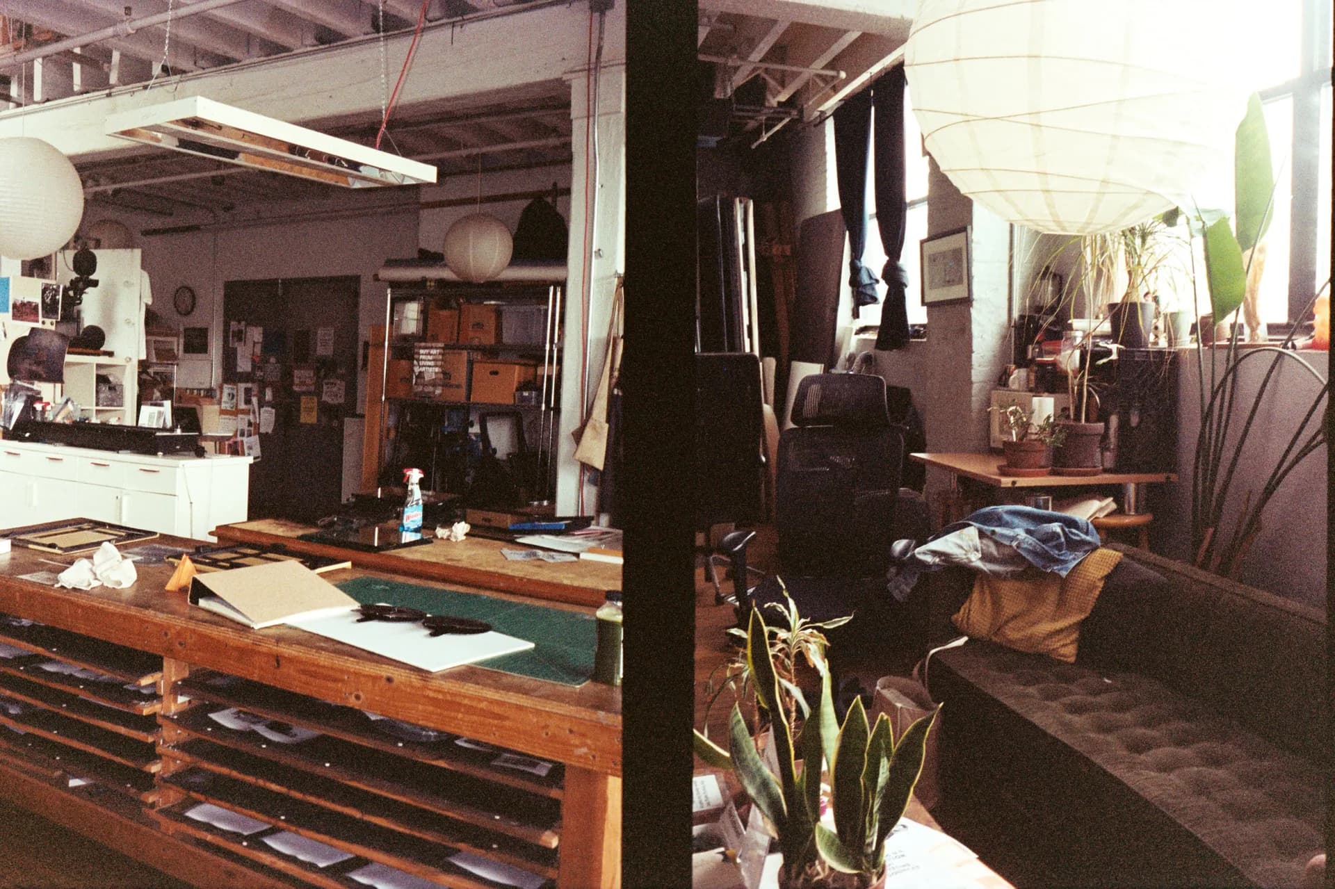 Interior of Gowanus Darkroom showing wooden work tables with contact sheets, plants, hanging prints, and darkroom workspace in natural daylight.