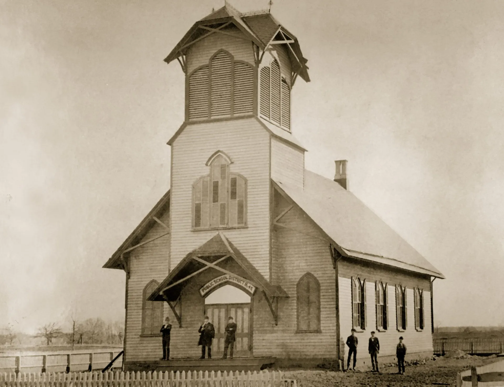 Sepia-toned photograph of a white wooden church with a tall steeple, white picket fence, and people standing outside on flat prairie landscape.