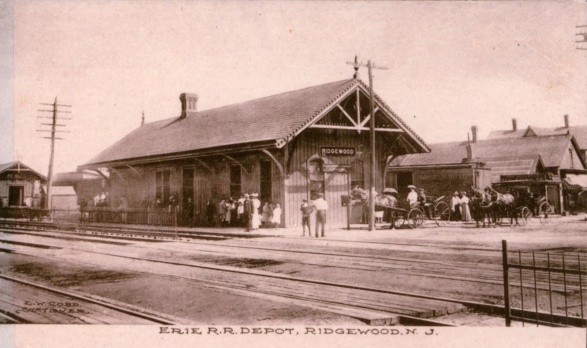 Hand-tinted photograph of Erie Railroad Depot in Ridgewood, New Jersey, showing wooden station building with people and horse-drawn carriages on tracks.