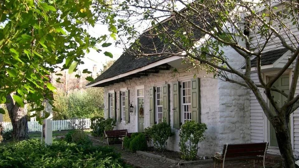 Exterior photograph of a small stone cottage with white trim, porch, shuttered windows, and surrounding vegetation.