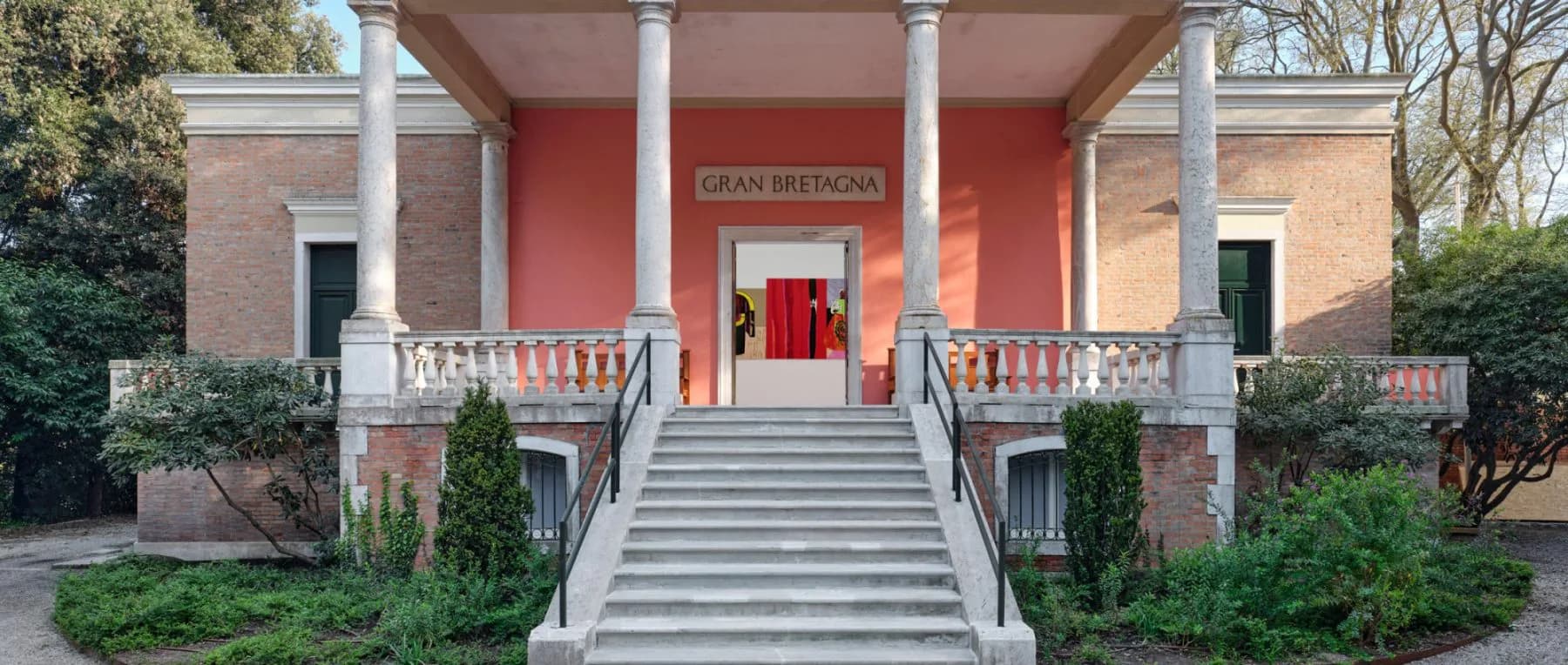Neoclassical brick building exterior with red entrance, white columns, balustrades, and centered staircase approach.