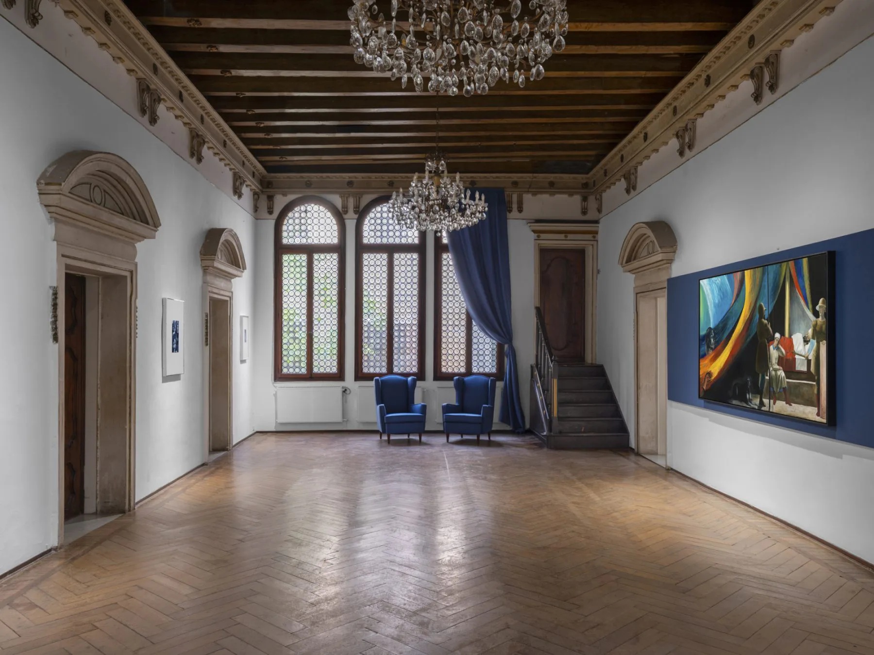 Elegant gallery interior with exposed wooden ceiling beams, crystal chandelier, tall arched windows, and contemporary blue artworks on white walls.