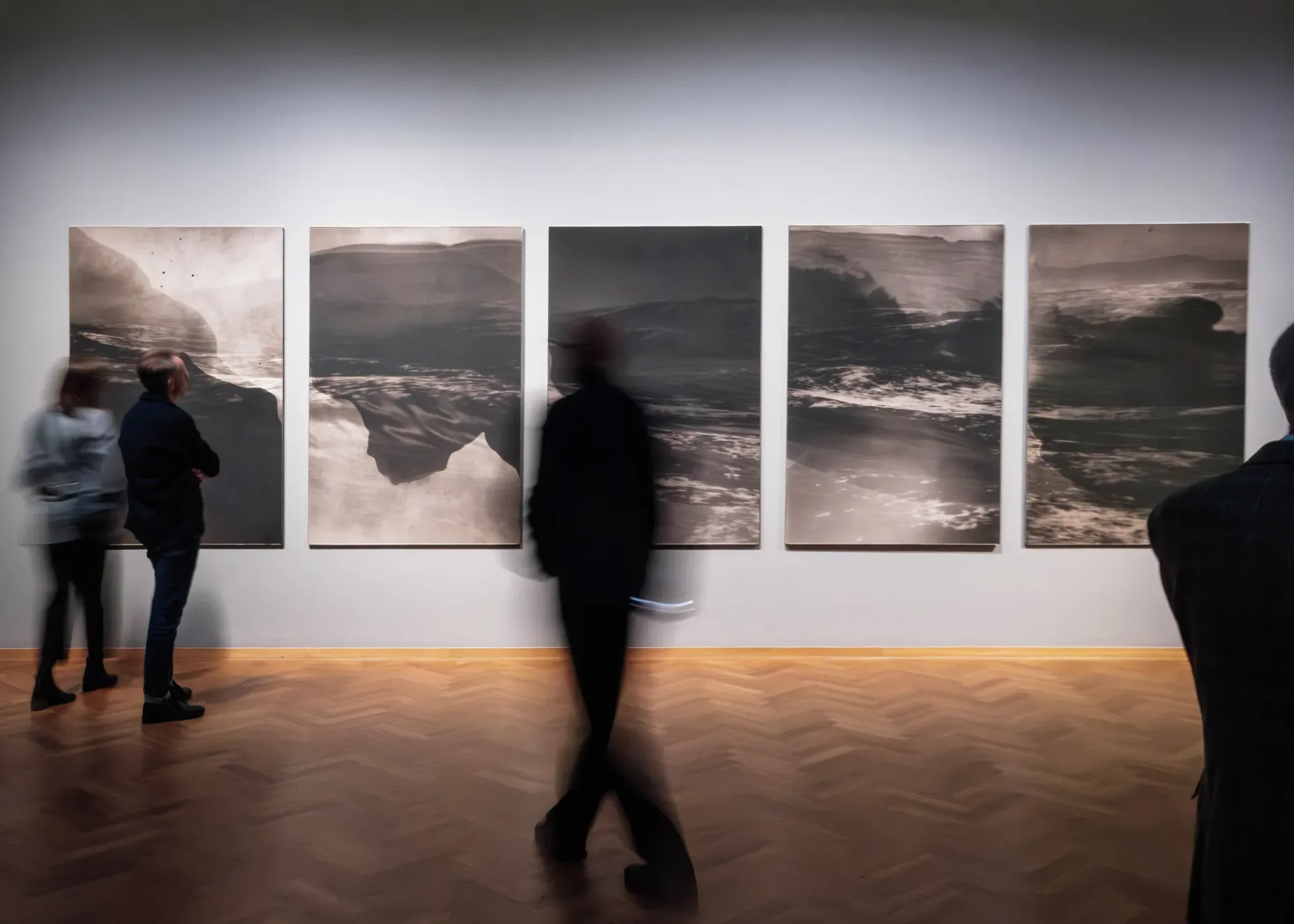 Gallery with dramatic lighting displaying five large-scale landscape photographs in neutral tones showing mountains and water, with visitors viewing the work.