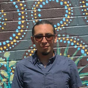 Headshot of person wearing glasses and blue patterned button-up shirt, posed in front of colorful dotted mural.