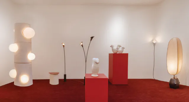 Gallery installation with red platform and white wall; white sculptural objects, lit vessels, and metallic stand displayed.