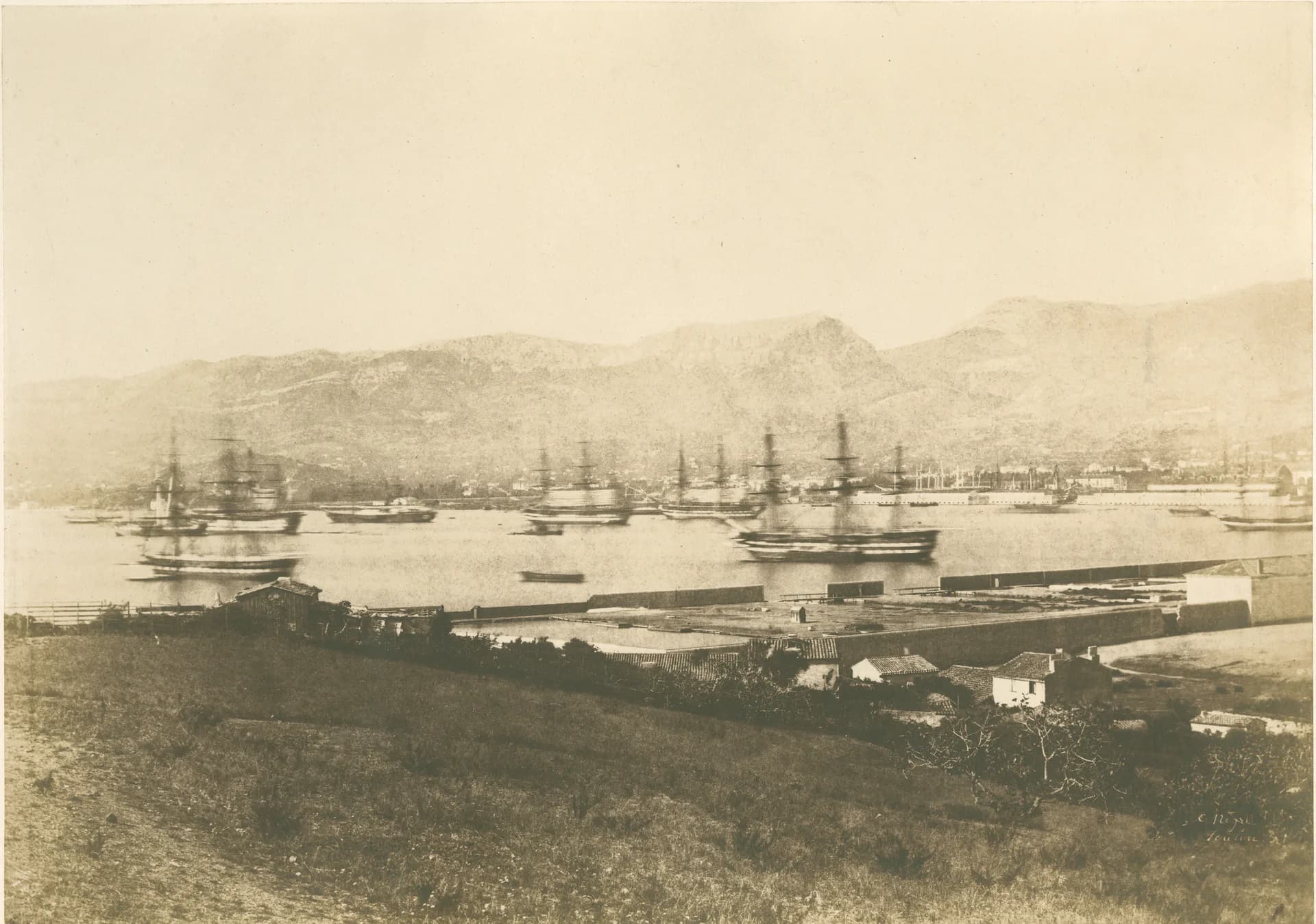 Sepia-toned photograph of a harbor with multiple sailing ships anchored in calm water, mountainous coastline beyond.