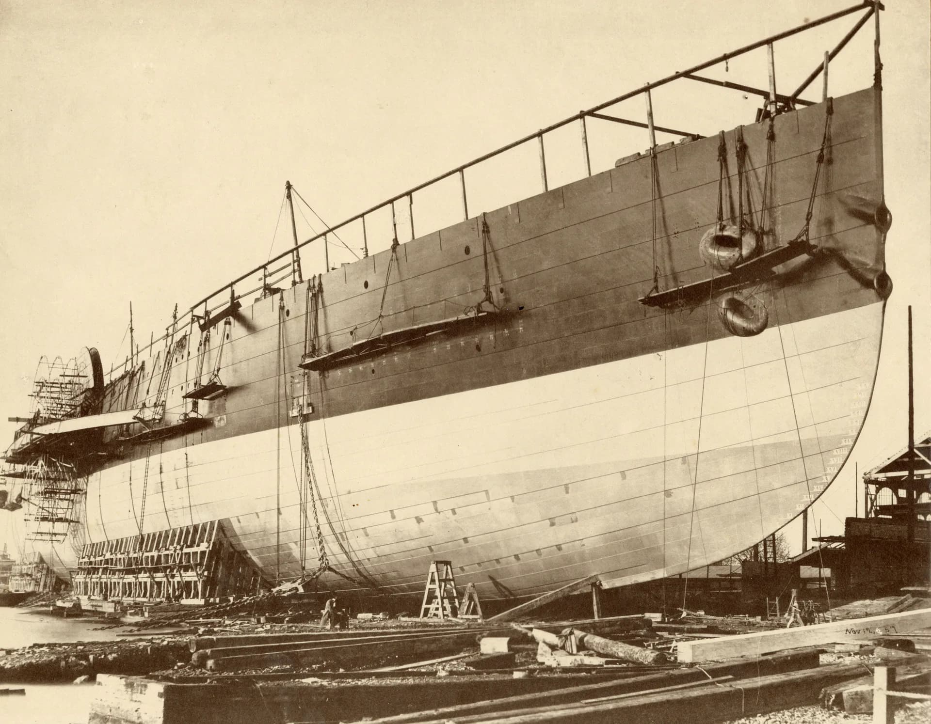 Sepia photograph of a large ship under construction in dry dock, showing wooden hull and metal framework overhead.