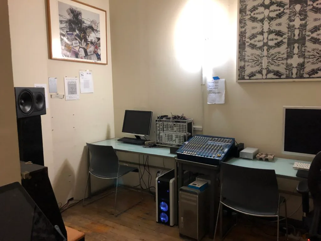 Harvestworks production workspace with mixing console, computer monitors, speakers, and equipment on desk under bright overhead light.
