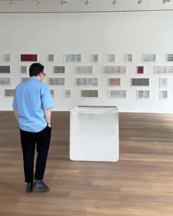 Gallery interior with white walls displaying a grid of framed artworks, a person in blue shirt viewing the wall-mounted collection.
