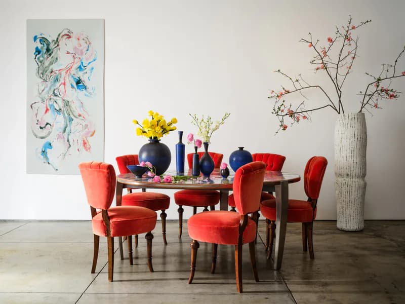 Styled gallery or showroom with red velvet chairs around dark table, watercolor artwork on wall, tall white vase with dried branches, dark flooring.