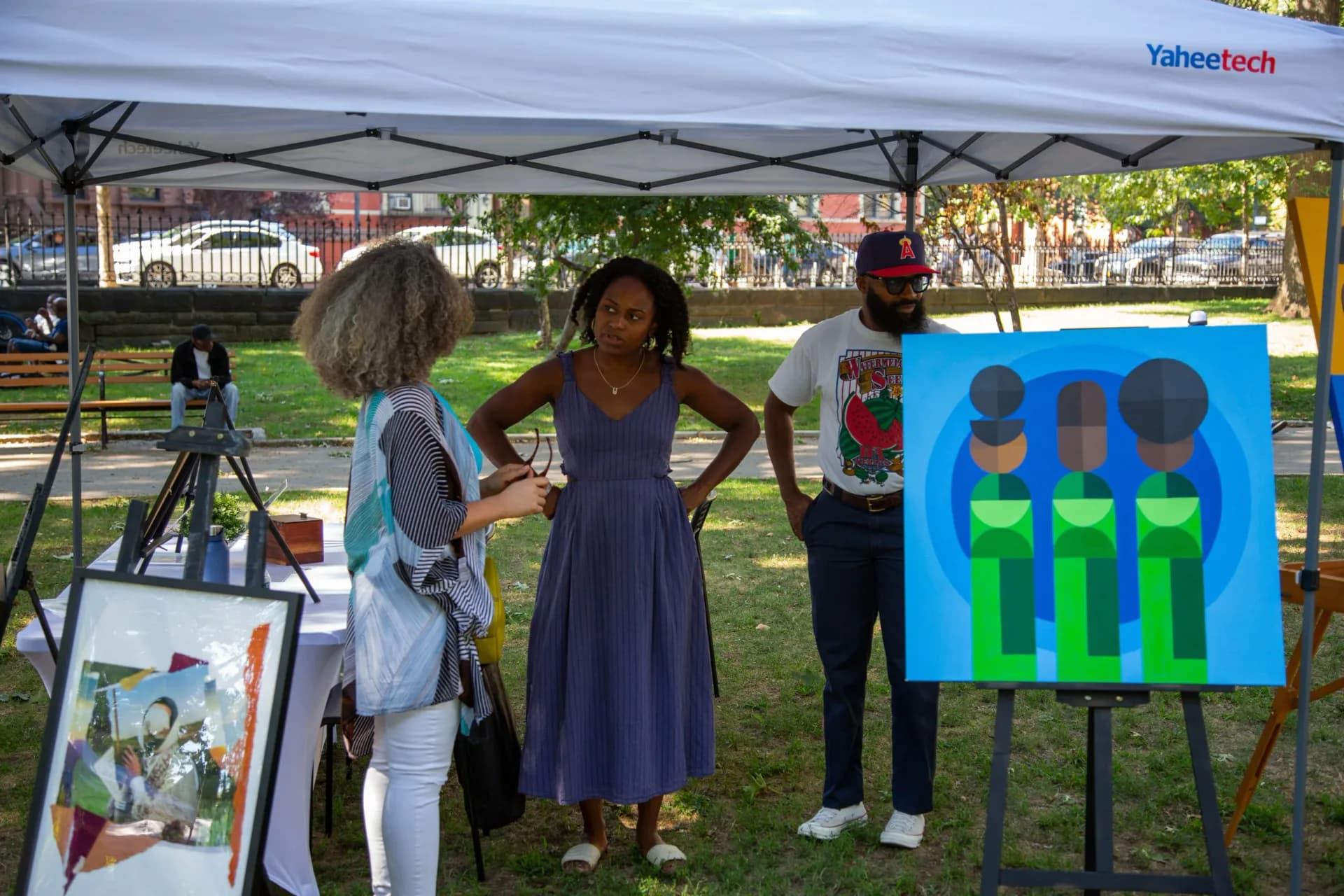 Heath Gallery pop-up booth at outdoor event with blue graphic artwork on display, visitors conversing with staff under white tent.